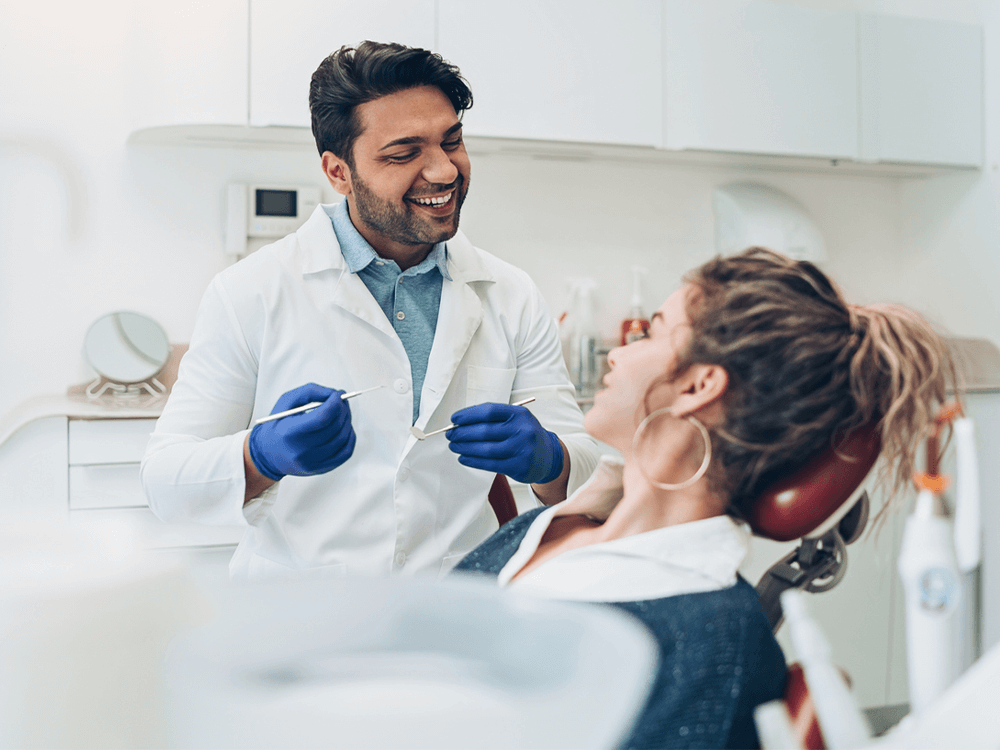 A friendly dentist wearing a white coat, blue gloves, and holding dental tools smiles while speaking with a female patient reclining in a dental chair. The modern dental office setting includes clean white cabinetry and equipment.