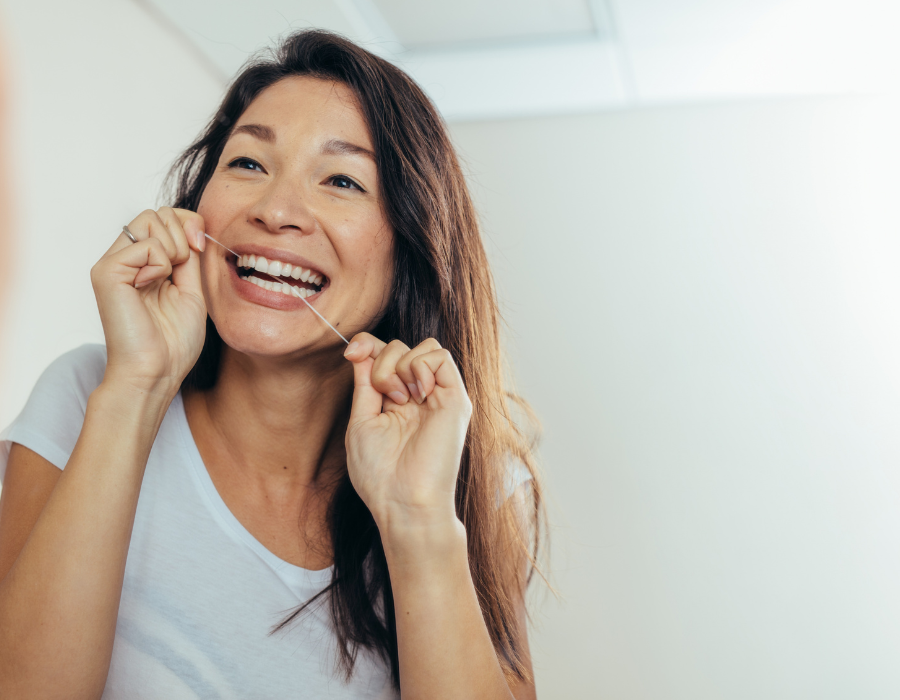 A smiling woman with long brown hair and a white shirt is flossing her teeth in a bright, clean setting. She maintains good oral hygiene with proper flossing technique.