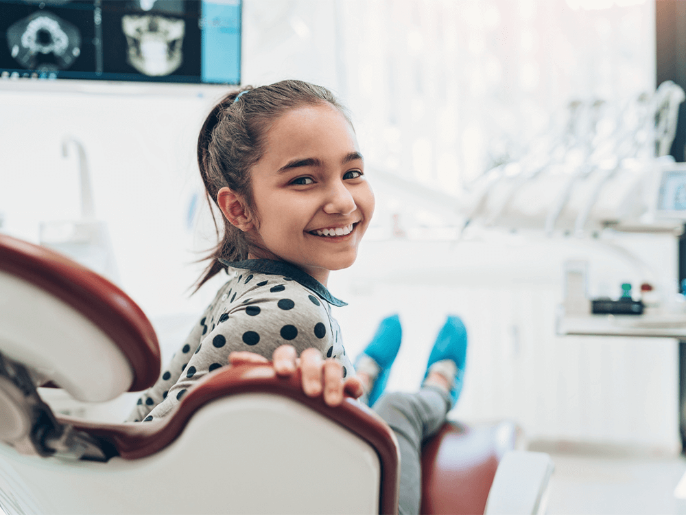 A happy young girl with a ponytail and a polka-dotted shirt sits in a dental chair, smiling at the camera. The bright, modern dental office features dental tools and a screen displaying X-ray images in the background.