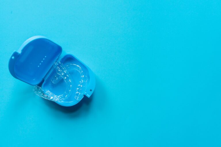 A pair of clear aligners placed inside an open blue protective case on a light blue background. The transparent trays are used for orthodontic treatment to straighten teeth.