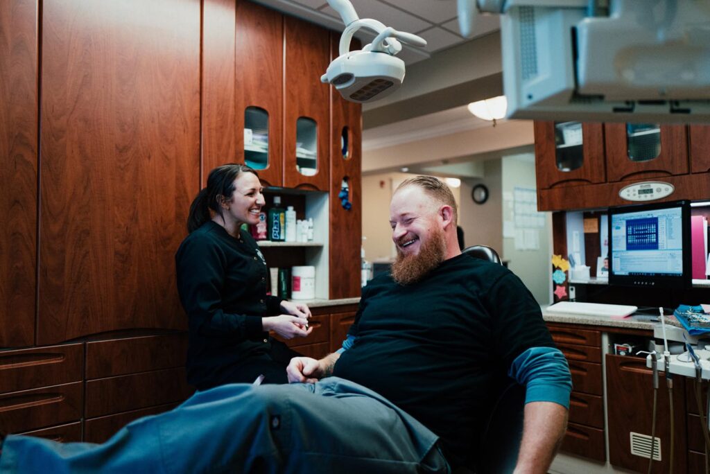 A bearded male patient reclines in a dental chair, smiling and laughing with a dental professional in a black uniform at Plaistow Dental. The office features wooden cabinetry, a dental light, and a computer displaying dental X-rays.