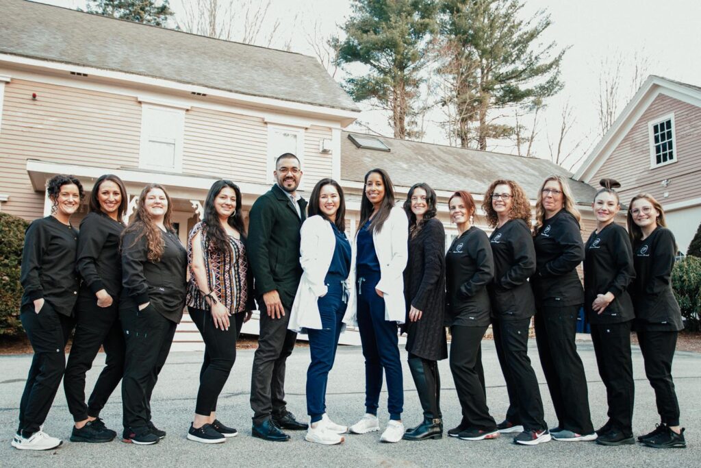 The Plaistow Dental team stands together outside their dental office, smiling for a group photo. The team includes dentists, hygienists, and staff members wearing professional attire, with some in lab coats and others in black uniforms.