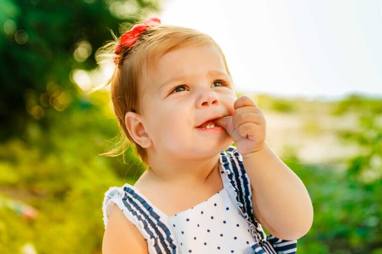 A smiling toddler with light brown hair and a red bow is outdoors in a sunlit setting, gently biting their finger. The child is wearing a white and navy polka-dotted outfit, showcasing emerging baby teeth.